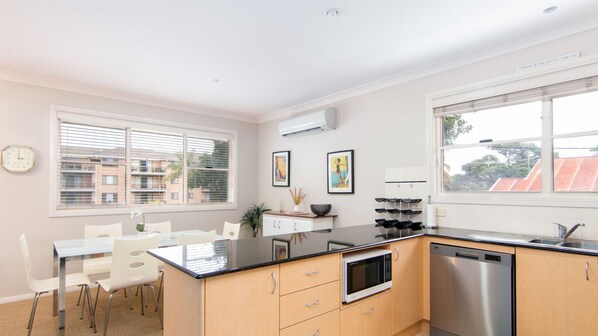 Cocina moderna con comedor, que incluye lavavajillas de acero inoxidable, microondas y pileta. Se observan cuadros enmarcados en la pared y un reloj.