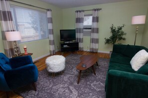 Olohuoneessa on sininen nojatuoli, vihreä sohva, puinen sohvapöytä, valkoinen ja pehmeä jalkarah, jalustalla oleva televisio ja ruukussa oleva viherkasvi.
