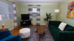 A living room with a green sofa, a blue chair, a white fluffy ottoman, a wooden coffee table, and a television on a black stand. - Art Inspired Cottage just 1 block from Main Street (Trinidad)