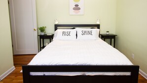 A neatly made bed with white bedding and two pillows with the words "RISE" and "SHINE" on them. There is a small plant on a bedside table and a wall art piece above the bed. - Art Inspired Cottage just 1 block from Main Street (Trinidad)