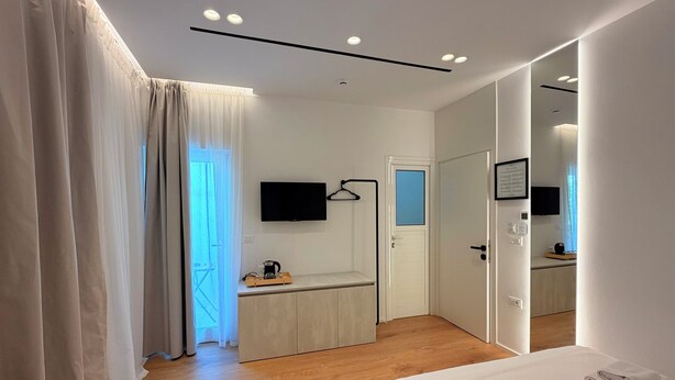 A modern hotel room with a bed, a desk with a flat-screen TV, a mirror, and a door leading to a bathroom. - Villa Mucho (Sarandë)