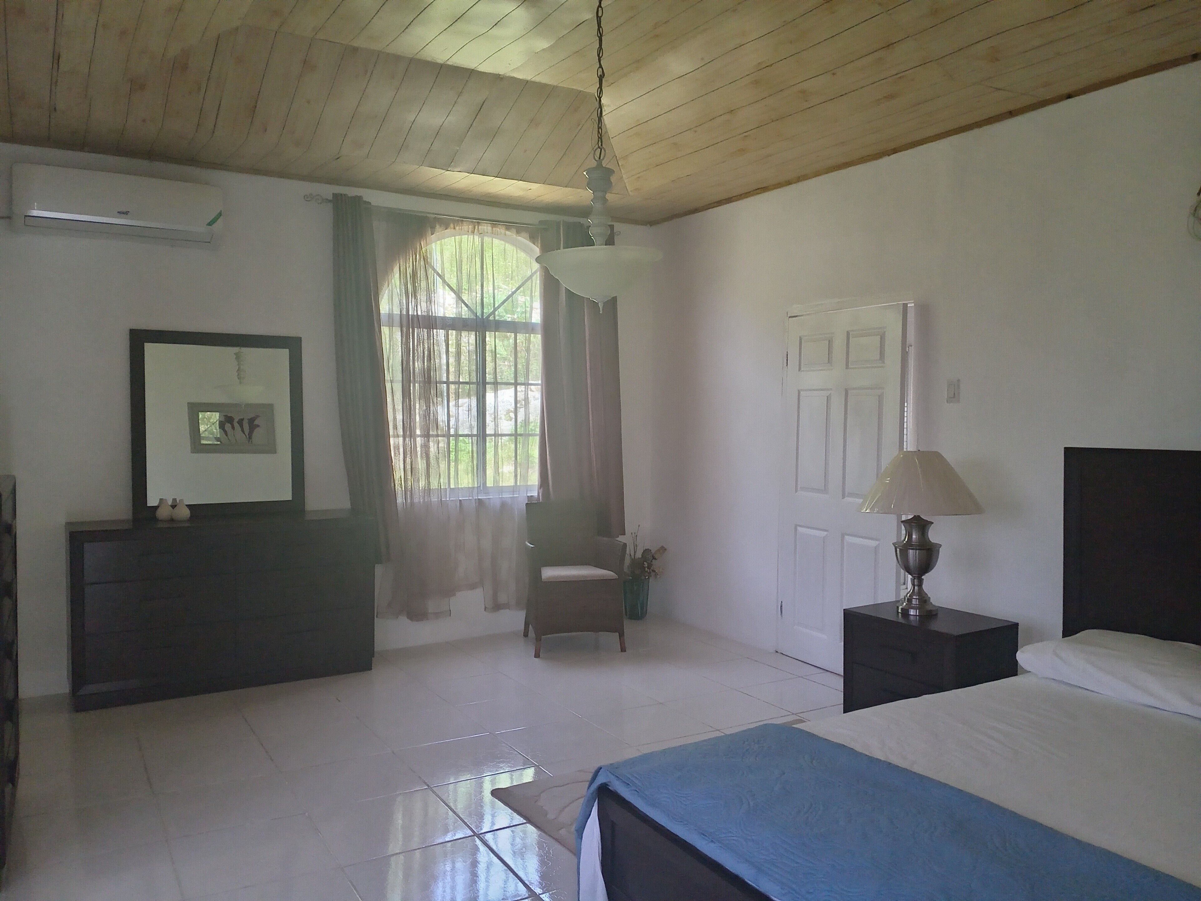 Un dormitorio con cama, mesita de noche, silla, ventana con cortinas, aire acondicionado y puerta.