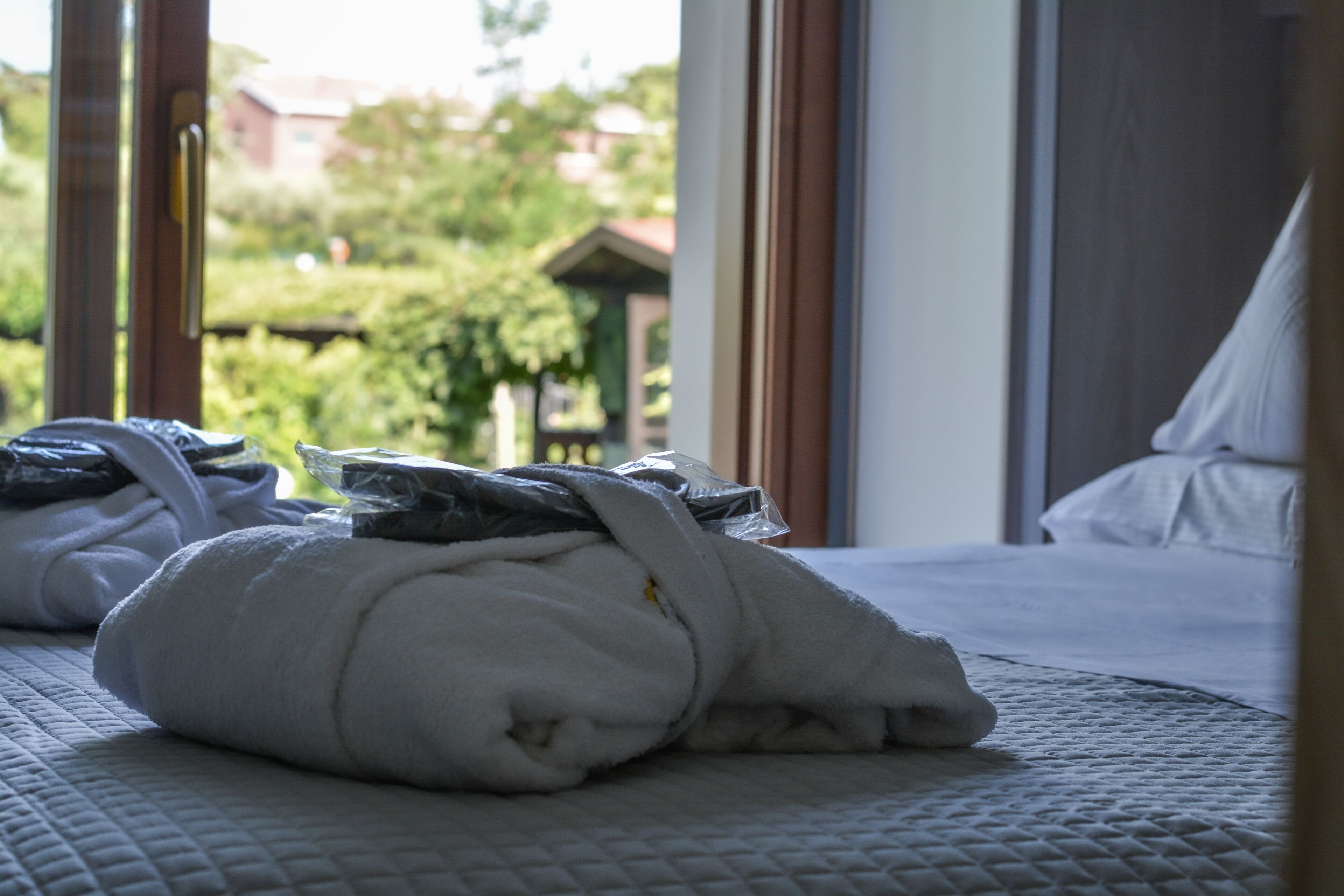A neatly made bed with a white duvet and rolled towels, a window with a view of greenery and buildings outside.