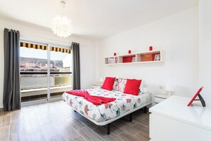 A bedroom with a bed, a bedside table, a nightstand, a shelf with decorative items, and a view through a large window. - Home2Book Beautiful Apart Los Cristianos Pool Wifi (Arona)