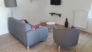Ein modernes Wohnzimmer mit grauem Sofa, einem grauen Sessel, einem kleinen runden Tisch, einer Stehlampe, einem an der Wand montierten Fernseher und einer Heizung.