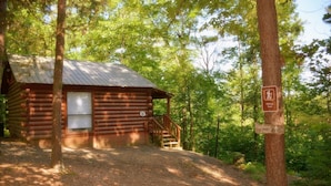 Une cabane rustique en rondins de bois, surmontée d’un toit en métal, avec un escalier en bois menant à une véranda et un panneau indiquant un sentier de randonnée à proximité.
