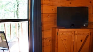 L’intérieur d’un chalet en bois, avec un téléviseur posé sur un meuble en bois, une fenêtre donnant sur les arbres et une chaise près de la fenêtre.