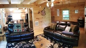 Een knus interieur van een chalet met een leren bank, een bijpassende fauteuil, een houten eettafel en een plafondventilator.