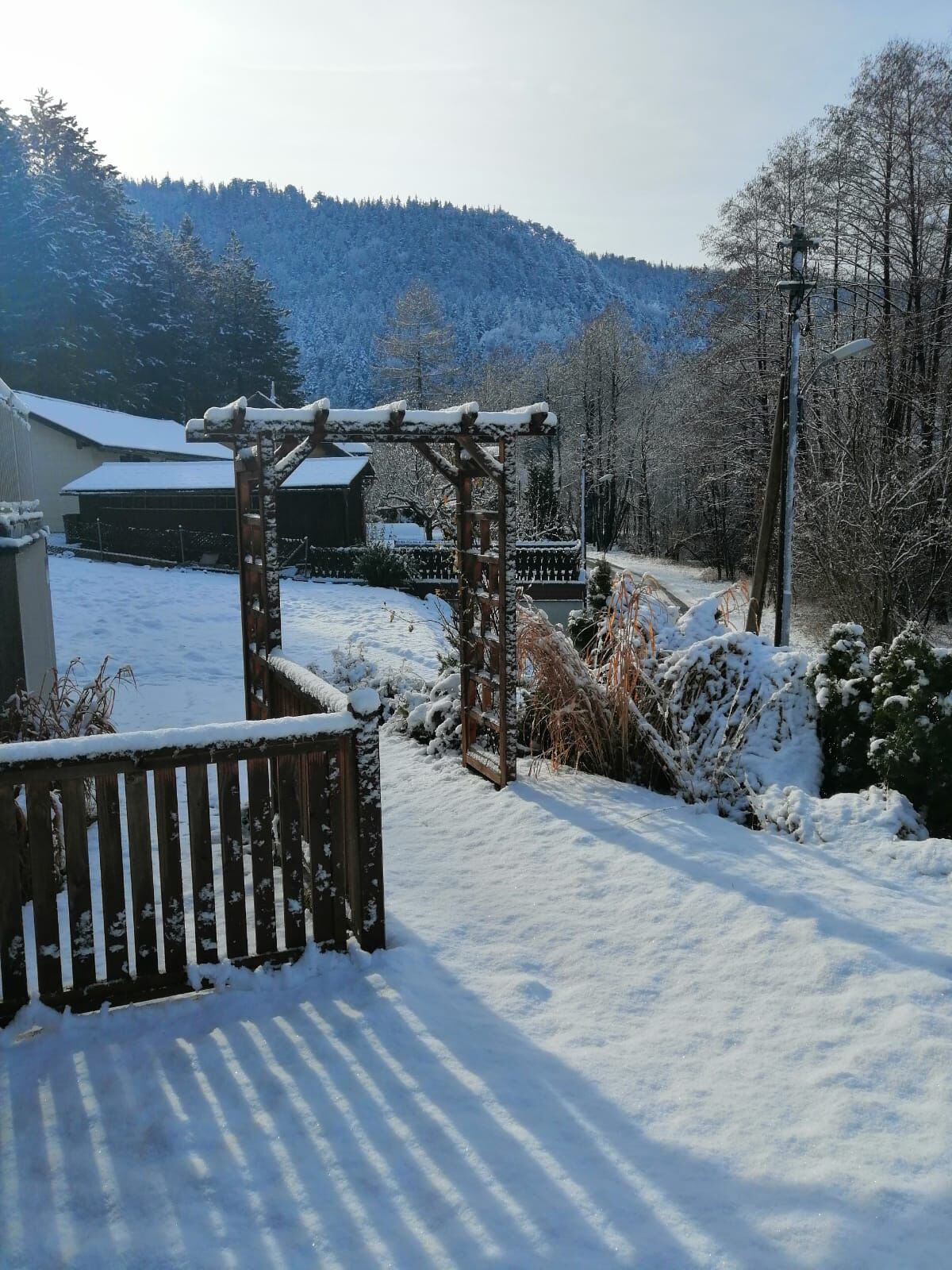 A snow-covered outdoor area with a wooden pergola, a bench, and a pathway leading to a building.