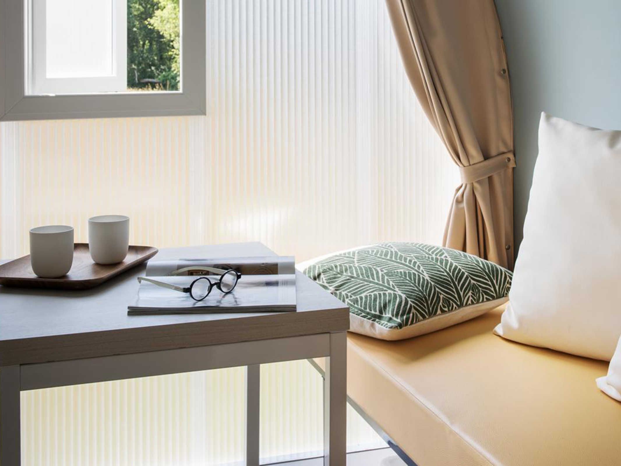 A room with a window, a table with cups and glasses, a pillow, and a bed.