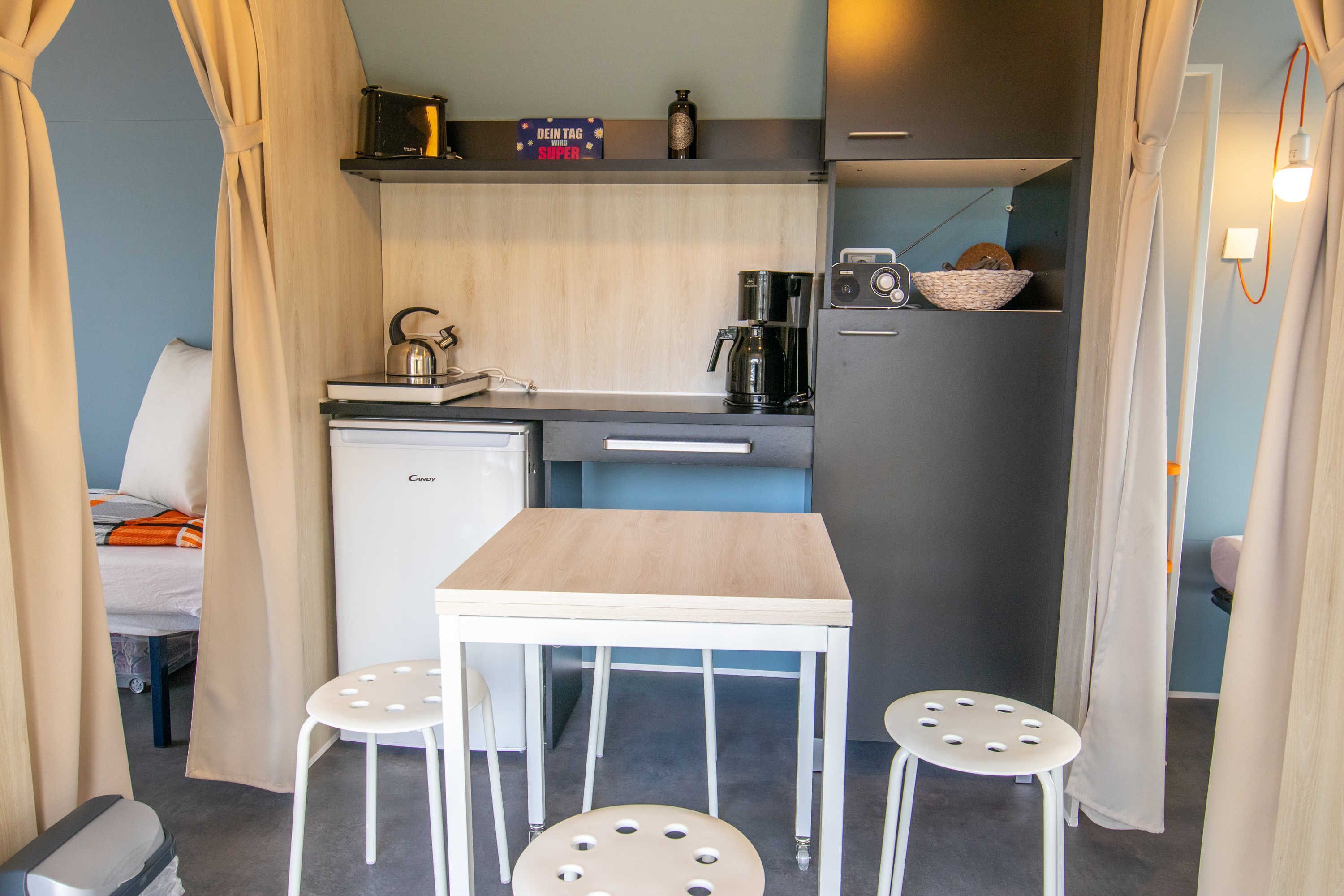 Una kitchenette compacta con cafetera, hervidor y radio. Hay una pequeña mesa con taburetes y una cama al fondo.