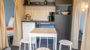 Uma cozinha compacta com máquina de café, chaleira e rádio. Há uma pequena mesa com banquetas e uma cama ao fundo.