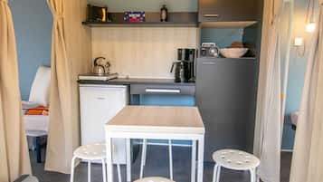 Una kitchenette compacta con cafetera, hervidor y radio. Hay una pequeña mesa con taburetes y una cama al fondo.