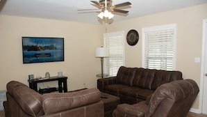 A living room with a brown leather sofa, a coffee table, and a ceiling fan. There is a painting on the wall and a clock on the wall. - About Legacy House @ Concord (Smyrna)