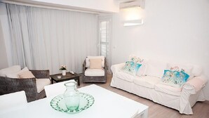 A living room with a white sofa, a wicker chair, a white table with a glass pitcher, and a window with curtains. - A very special apartment for up to 5 people. (Colònia de Sant Jordi)