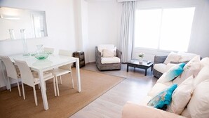 A bright living room with a white sofa, a dining table with chairs, a small side table, and a window with curtains. - A very special apartment for up to 5 people. (Colònia de Sant Jordi)