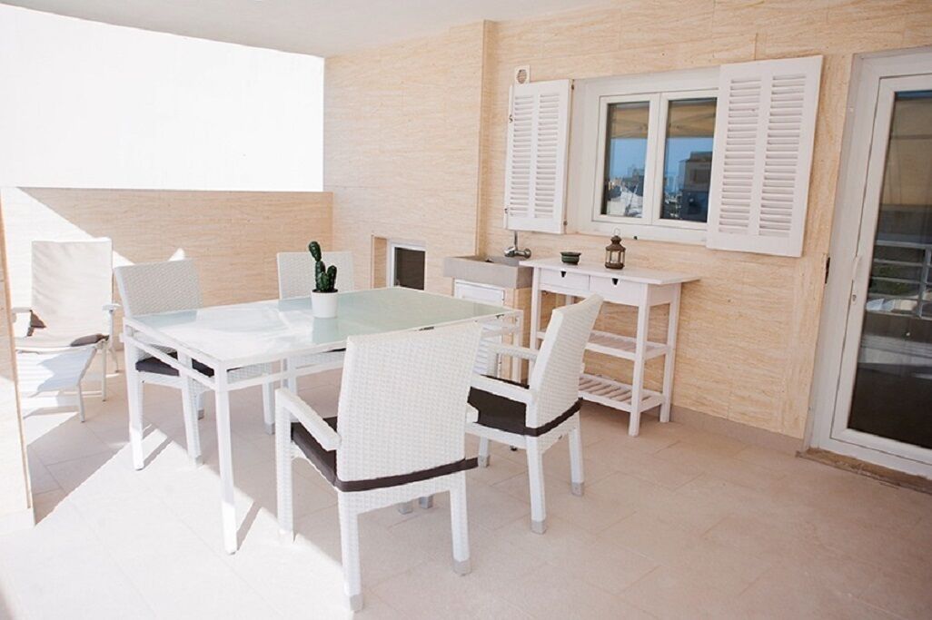 Een zonnig, licht overgoten terras met een witte tafel en stoelen, een kleine cactusplant en een raam met luiken.
