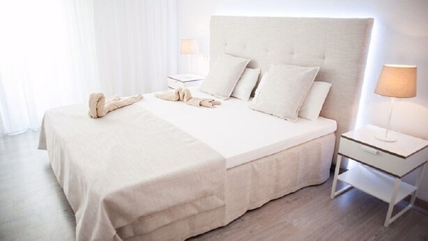 A neatly made bed with a white headboard, two bedside tables with lamps, and a window with sheer curtains. - A very special apartment for up to 5 people. (Colònia de Sant Jordi)