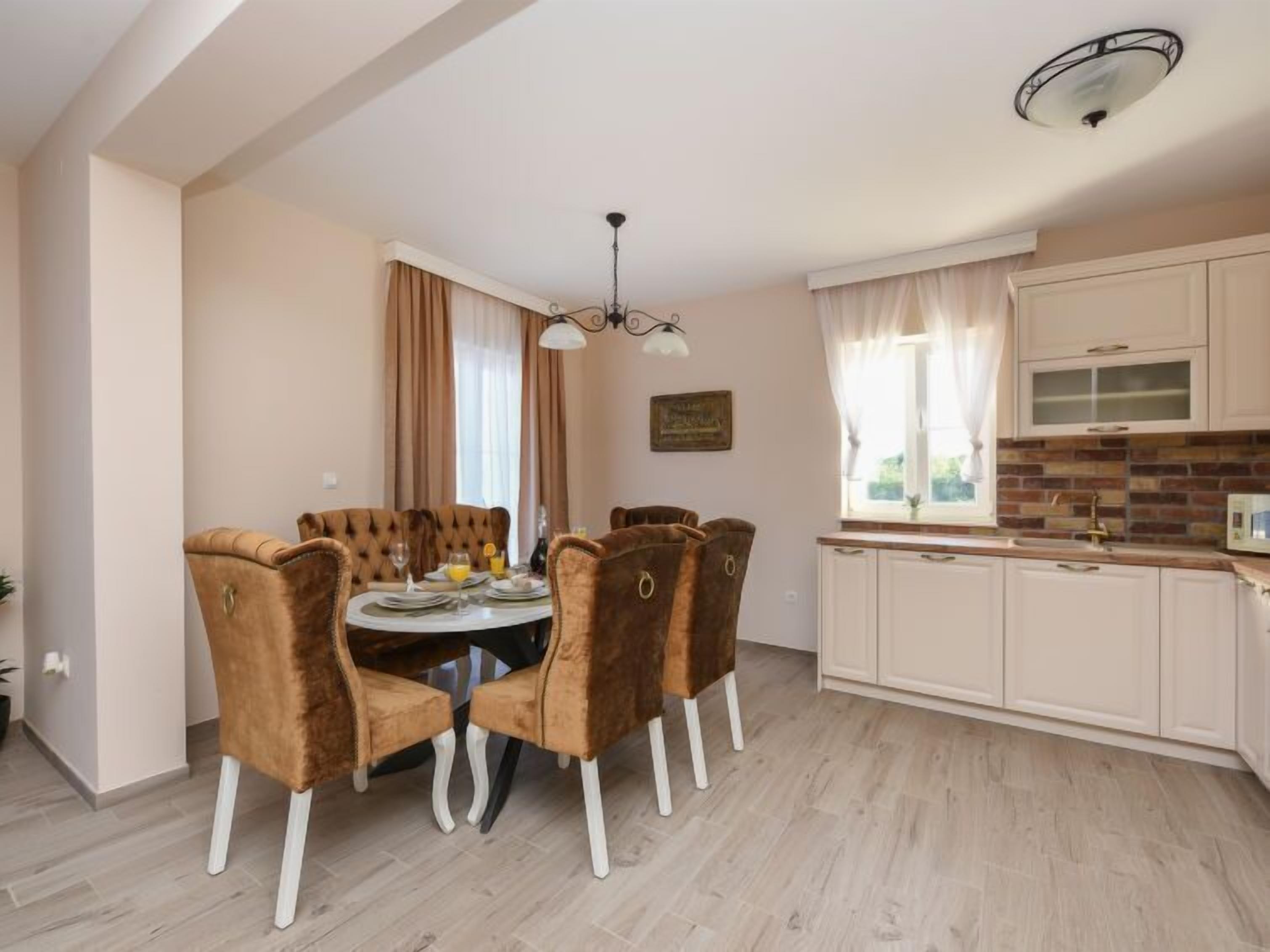 A dining area with a round table and six brown chairs, a kitchen with white cabinets, and a window with curtains.