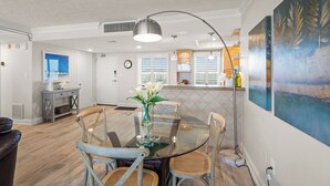 Une cuisine moderne avec des meubles blancs, un coin repas doté d’une table en verre et de chaises en bois, et un grand tableau accroché au mur.