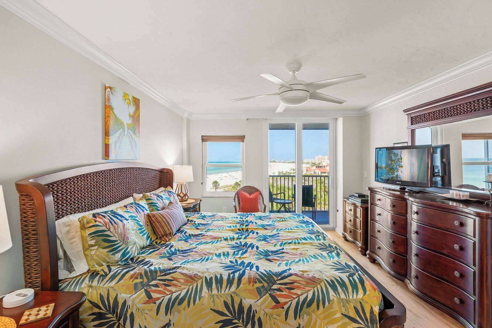 A bedroom with a bed, a ceiling fan, a TV, a dresser, and a balcony with a view of the beach.