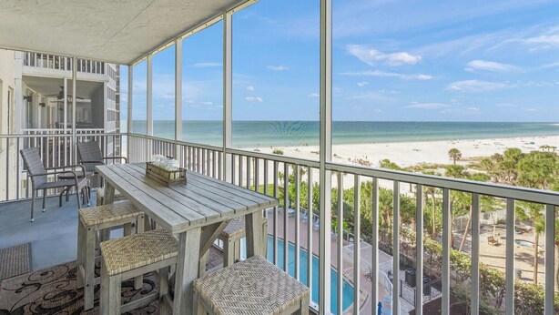 Un balcon avec une table en bois et des chaises en osier, donnant sur une plage sous un ciel dégagé.
