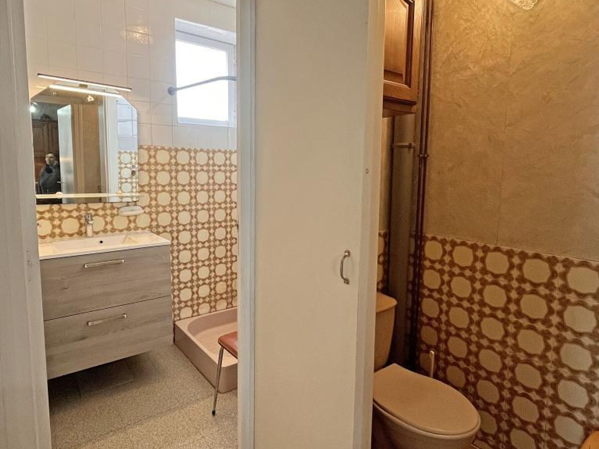 Een badkamer met een behang, een toilet, een wastafel met spiegel en een klein bankje.