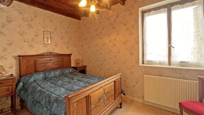 Un dormitorio con cama de madera, mesita de noche, cómoda, ventana con cortinas de encaje y radiador.