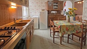 Una cocina con armarios de madera, una estufa a gas, un lavamanos y un refrigerador. Hay una mesa de comedor con sillas y un florero con flores.