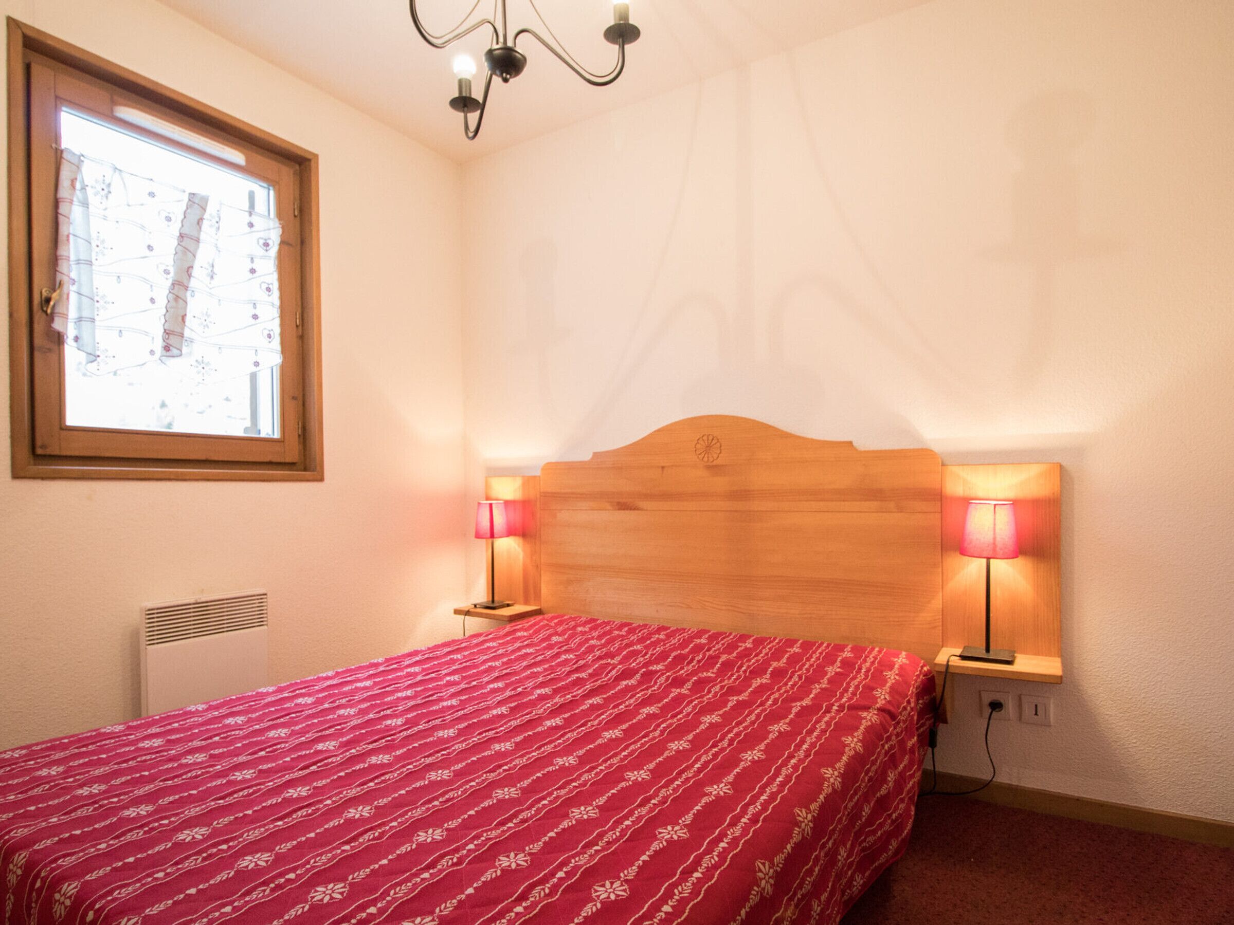 A bedroom with a wooden headboard, two bedside lamps, a red patterned bedspread, a window with blinds, and a radiator.