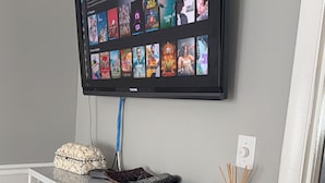 Een flatscreen-tv hangt aan de muur en toont een selectie films. Daaronder bevindt zich een witte plank met verschillende items, waaronder een rieten mand, afstandsbedieningen en boeken.