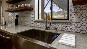 A kitchen with a stainless steel sink, wooden shelves, and a window with a view of mountains. - Joshua Tree Desert Retreat (Joshua Tree)