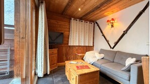 Interior acogedor de una cabaña con techo de madera, un sofá, una mesa de centro de madera, un televisor y una ventana con cortinas.