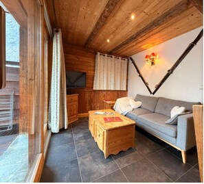 A cozy cabin interior with a wooden ceiling, a sofa, a wooden coffee table, a TV, and a window with curtains.