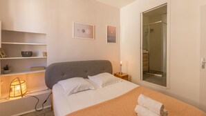 Un dormitorio con cama, mesitas de noche, lámpara, estante con objetos decorativos y ventana con vista a la zona de ducha.