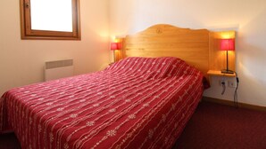 Ein Schlafzimmer mit einem hölzernen Kopfteil, einer rot gemusterten Bettdecke, zwei Nachttischlampen, einem Fenster und einer Heizkörper.