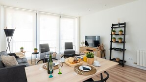 En moderne stue med spiseplads, store vinduer, et fladskærms-tv og en sort reol med planter.