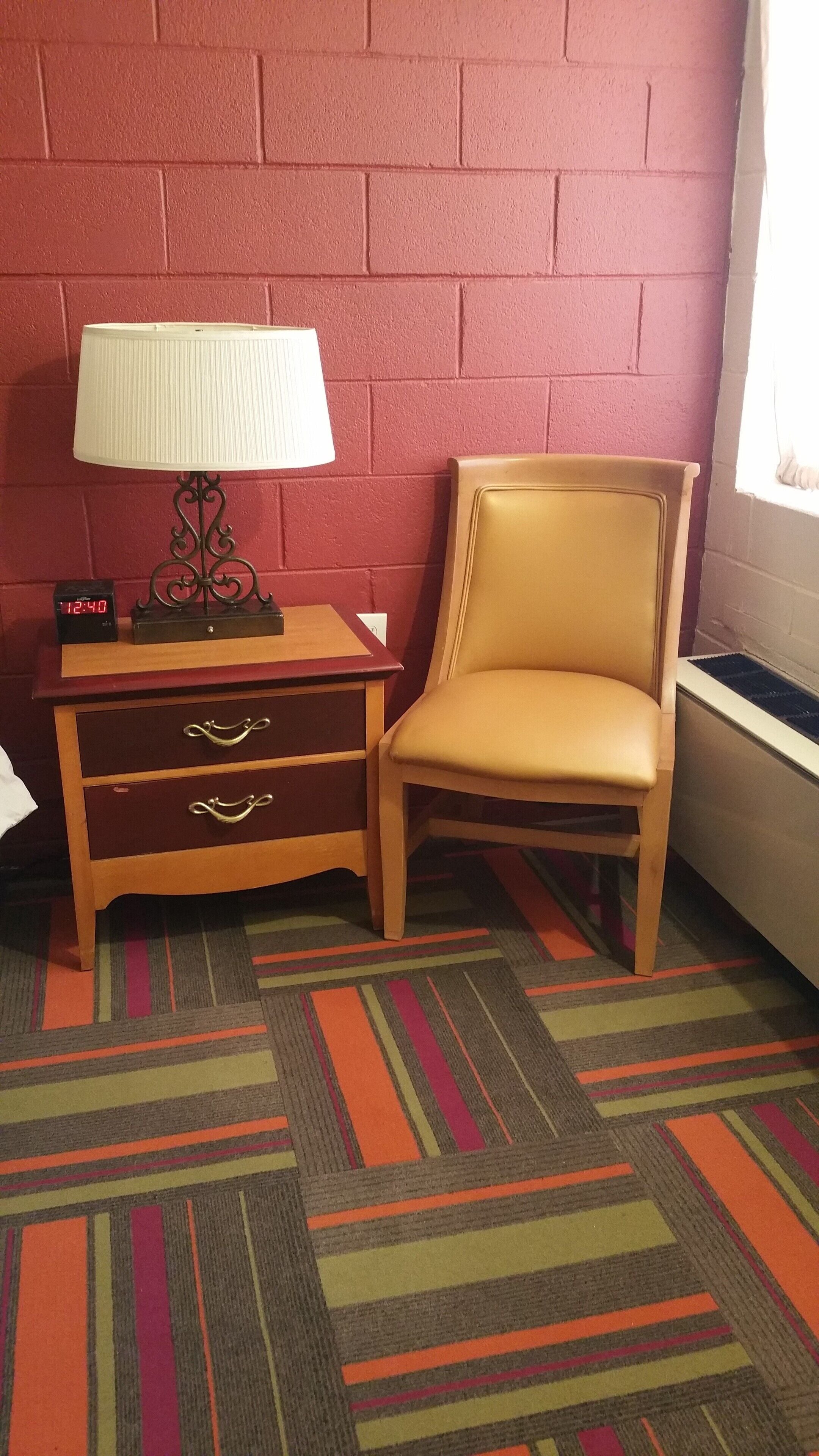 A room with a red brick wall, a wooden nightstand with a lamp, a chair, and a clock.