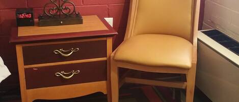 A room with a red brick wall, a wooden nightstand with a lamp, a chair, and a clock.