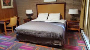 A hotel room with a brick wall, a bed with a headboard, two bedside tables with lamps, a wooden desk, a chair, and a colorful carpet. - Thunder Bird Motel (Mountain Home)
