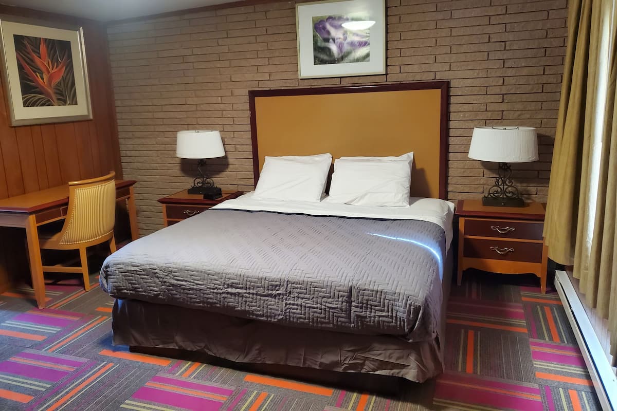 A hotel room with a brick wall, a bed with a headboard, two bedside tables with lamps, a wooden desk, a chair, and a colorful carpet.