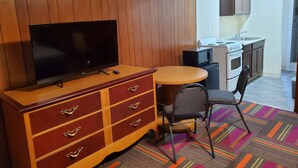 A compact room with a wooden panel wall, a flat-screen TV on a wooden cabinet, a small round table with a chair, a microwave, and a stove in the background. - Thunder Bird Motel (Mountain Home)
