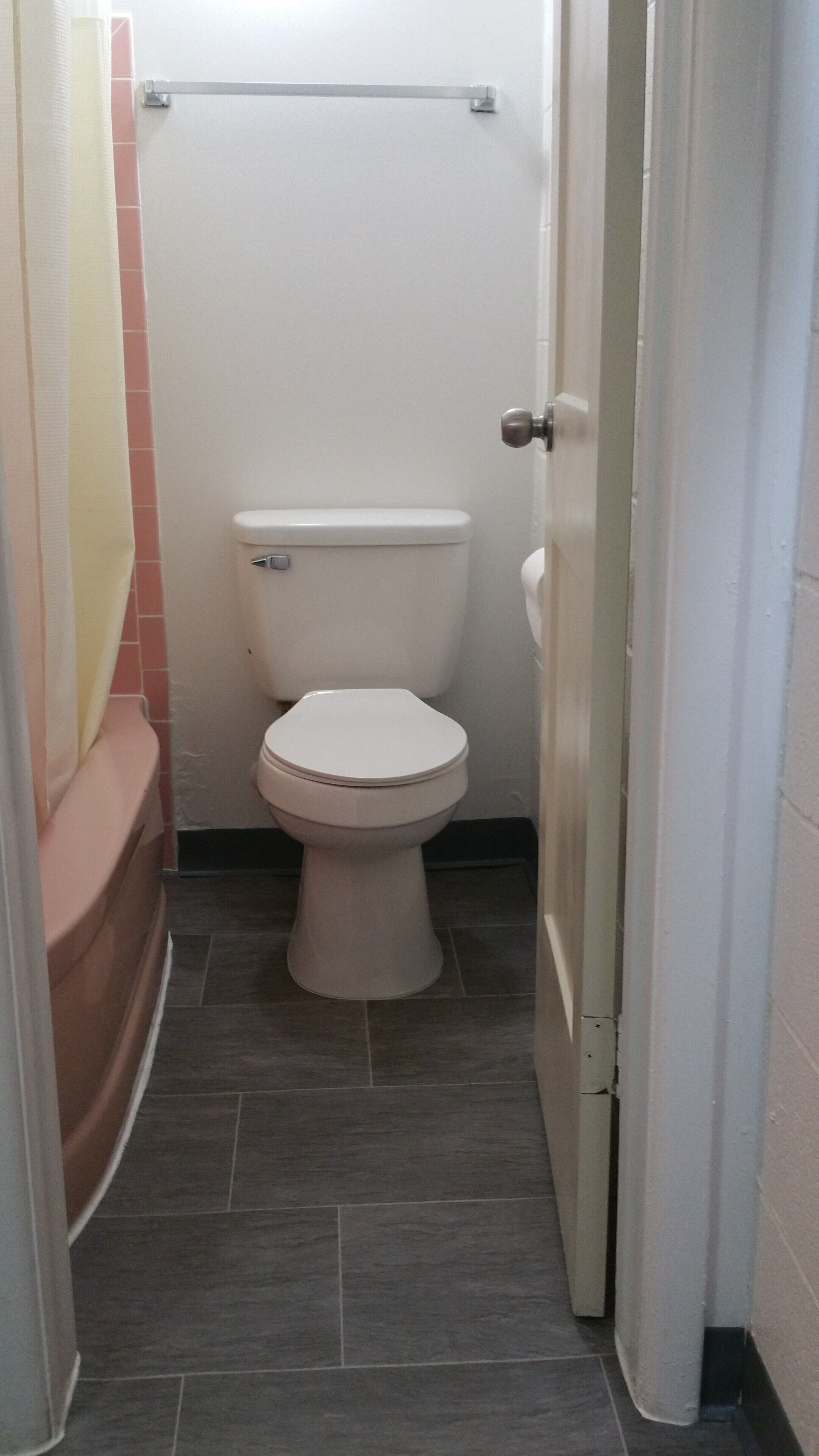 A small, narrow bathroom with a white toilet, a pink bathtub, and a tiled floor.