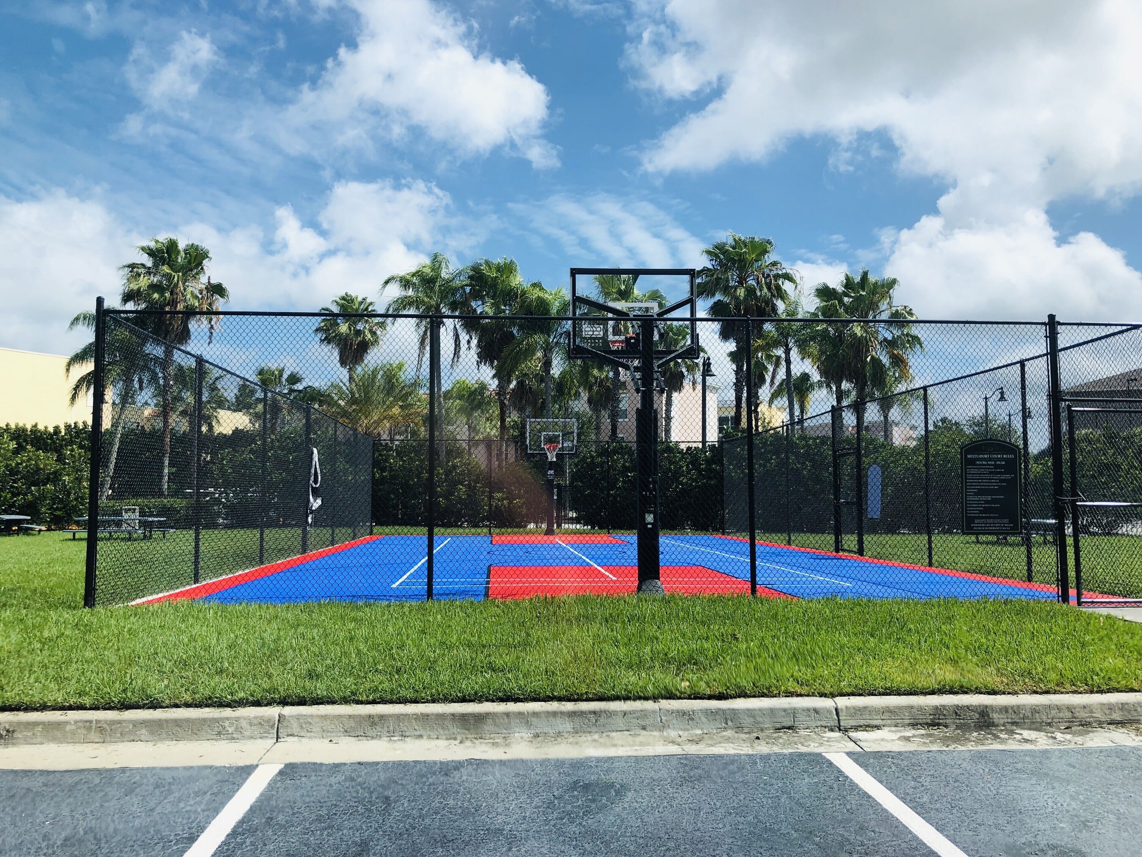 Un terrain de basket entouré d’une clôture en treillis métallique, avec des palmiers et un ciel dégagé.