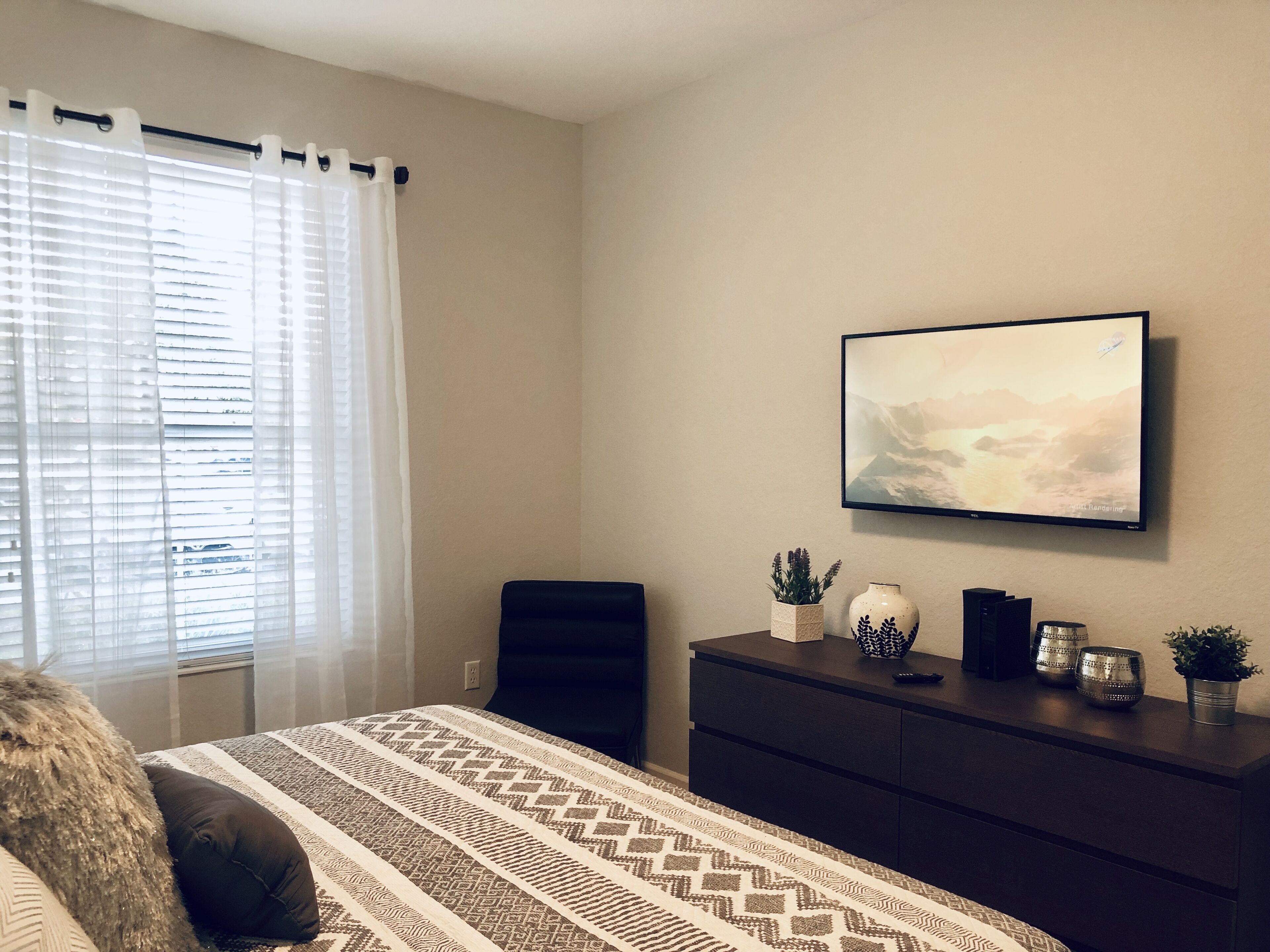 Une chambre à coucher avec un lit, une commode, un téléviseur fixé au mur et une fenêtre avec des rideaux.