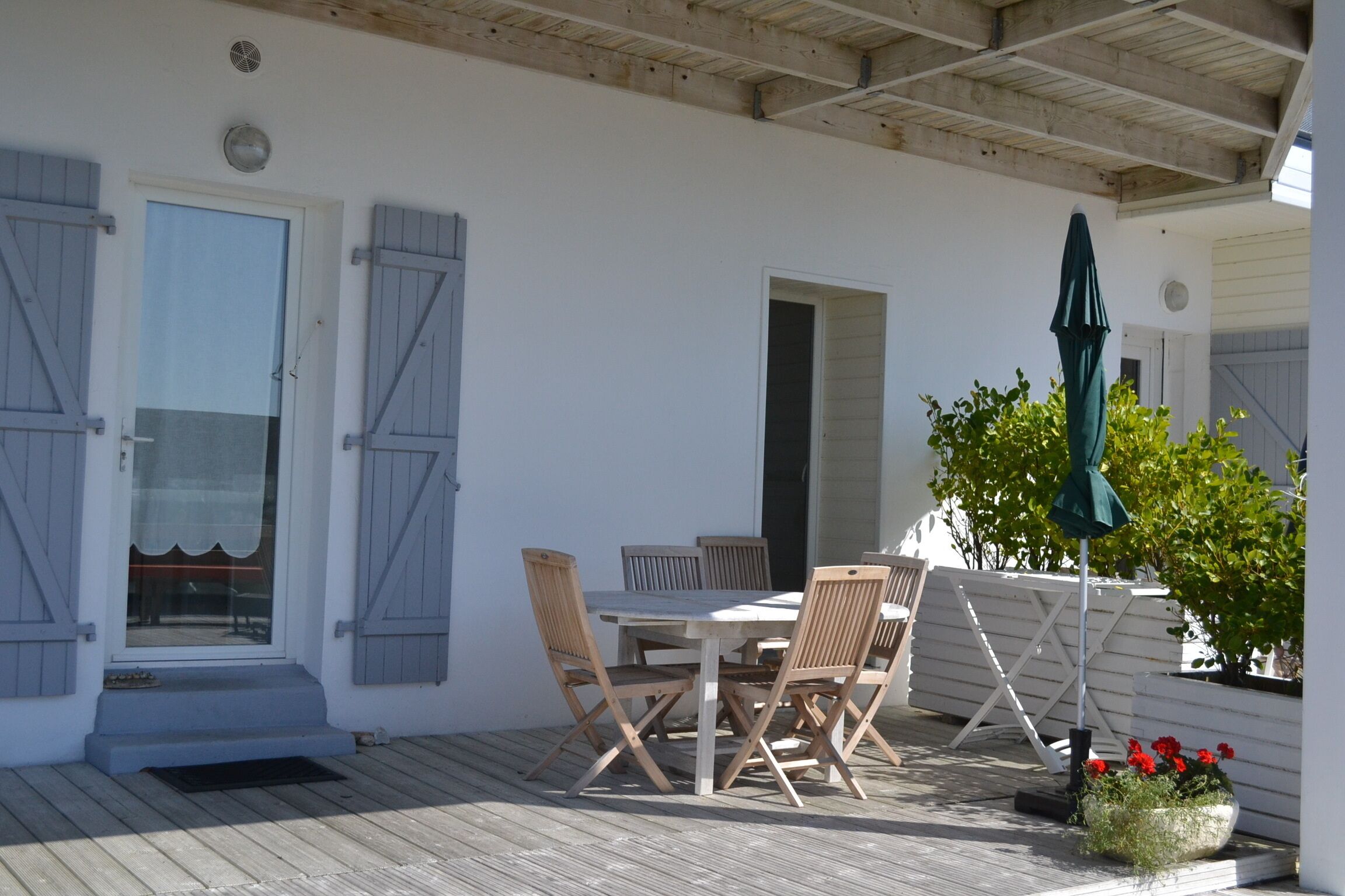 Un patio techado con mesa y sillas de madera, una puerta con contraventanas y una maceta con flores rojas.