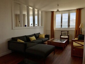 A living room with a sectional sofa, a glass coffee table, a wooden chair, and a view of the sea through large windows.