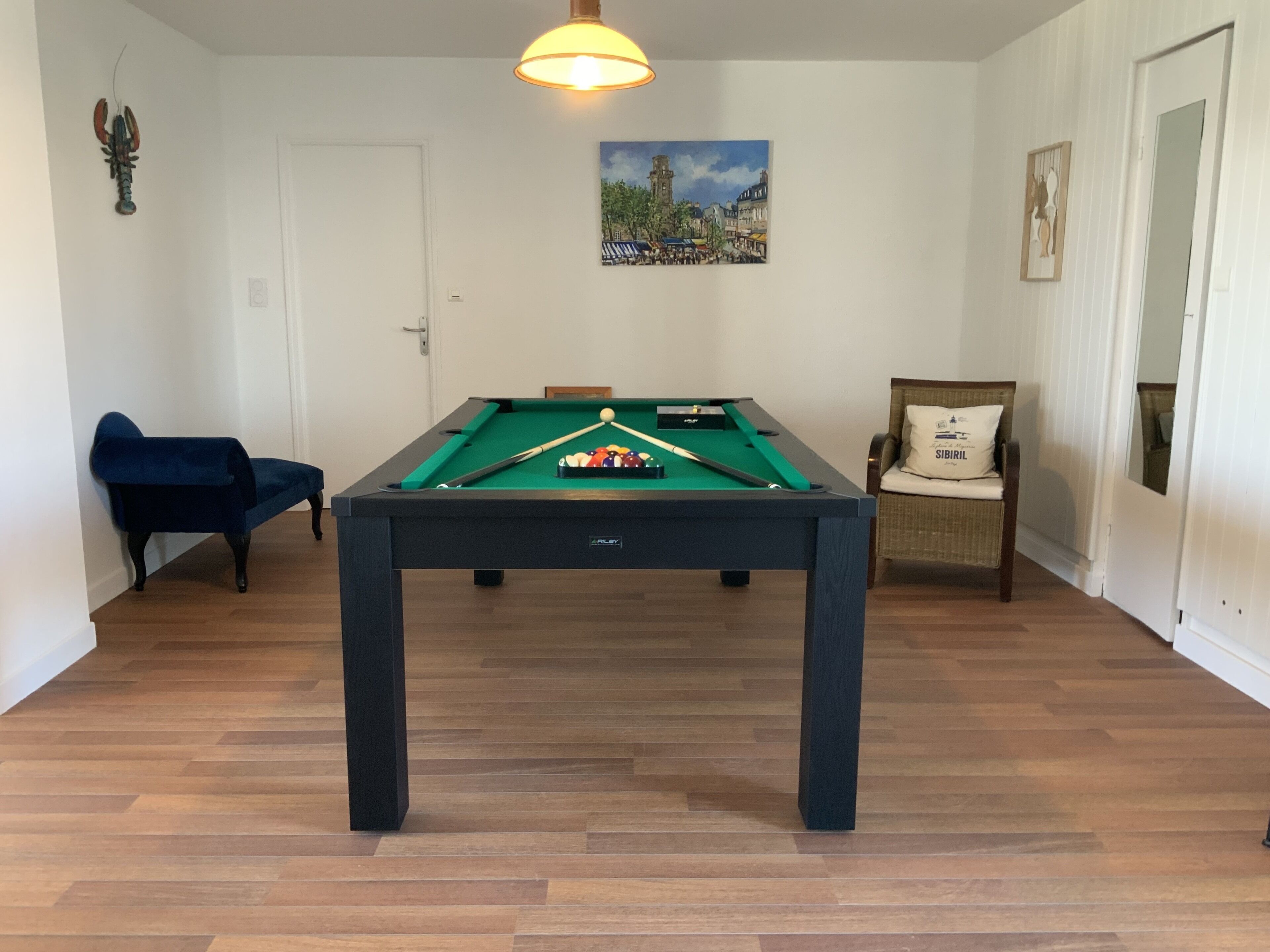 A pool table in a room with wooden flooring, a blue chair, a cushioned chair, and a painting on the wall.