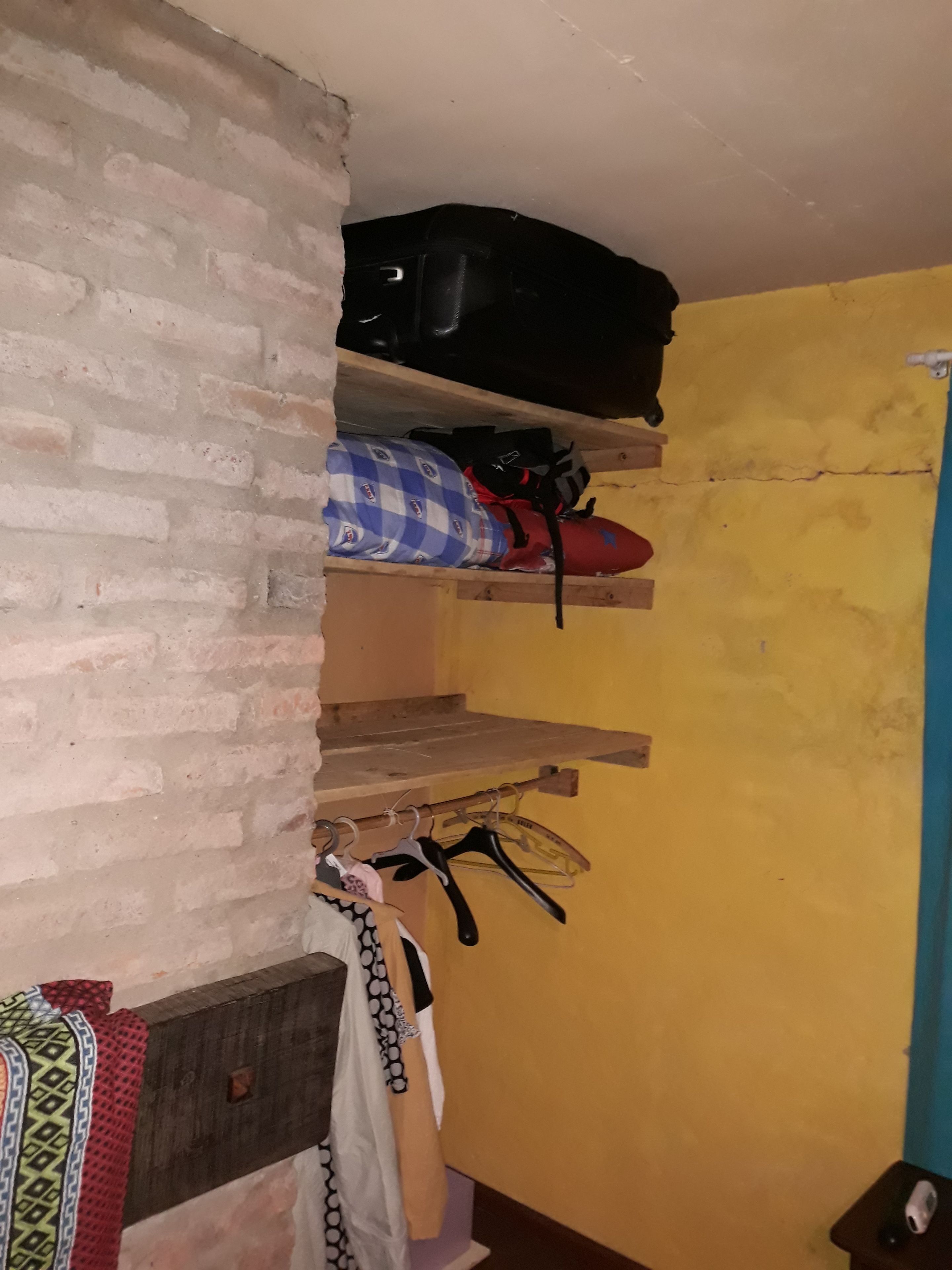 A room with a brick wall, a wooden shelf with a black suitcase, a blue and red bag, and a hanging coat rack with various items.
