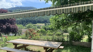 Une table de pique-nique en bois, avec des bancs, installée dans un espace extérieur verdoyant, entourée d’arbres et offrant une vue sur un paysage rural avec des maisons et des collines en arrière-plan.